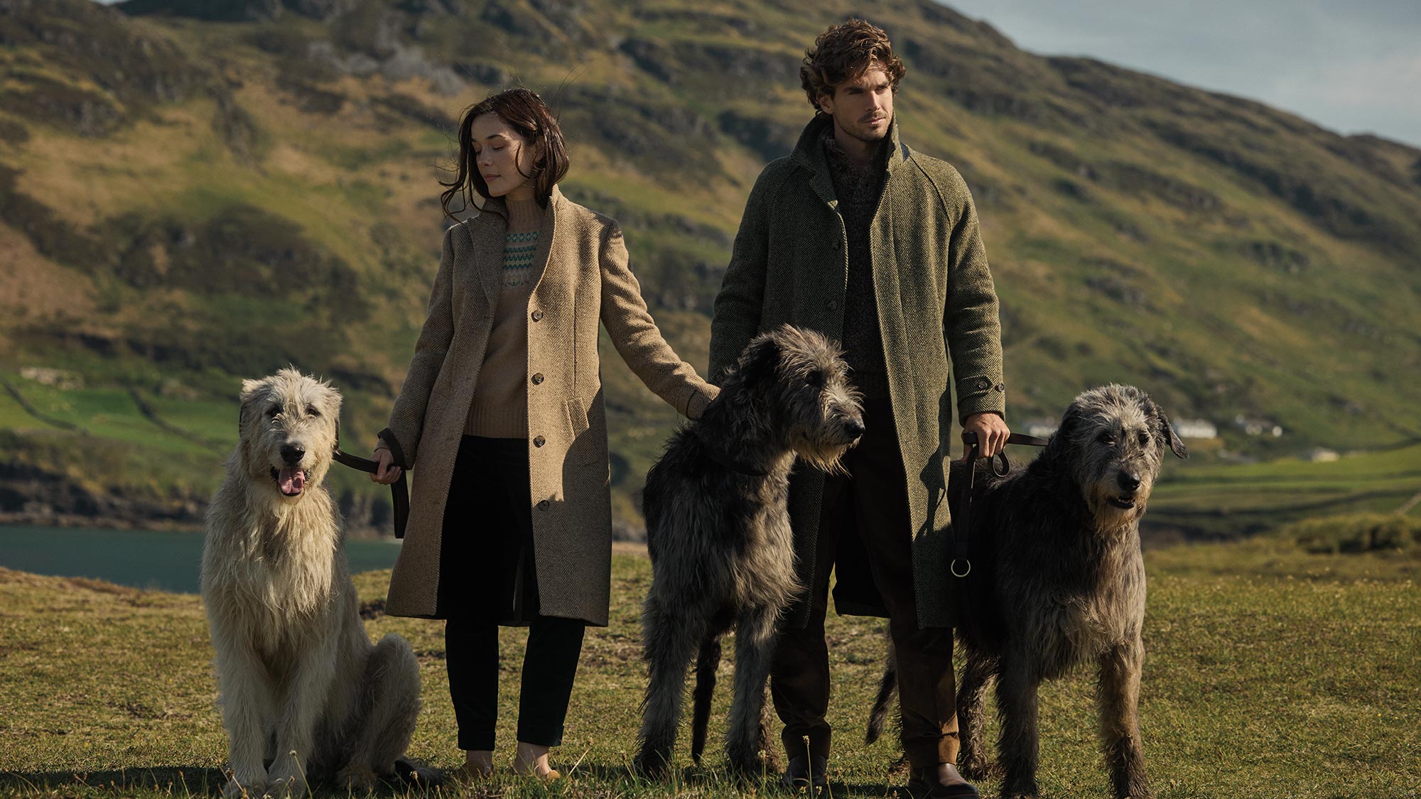 A man and woman stand on grassy terrain with three large dogs, mountains in the background.