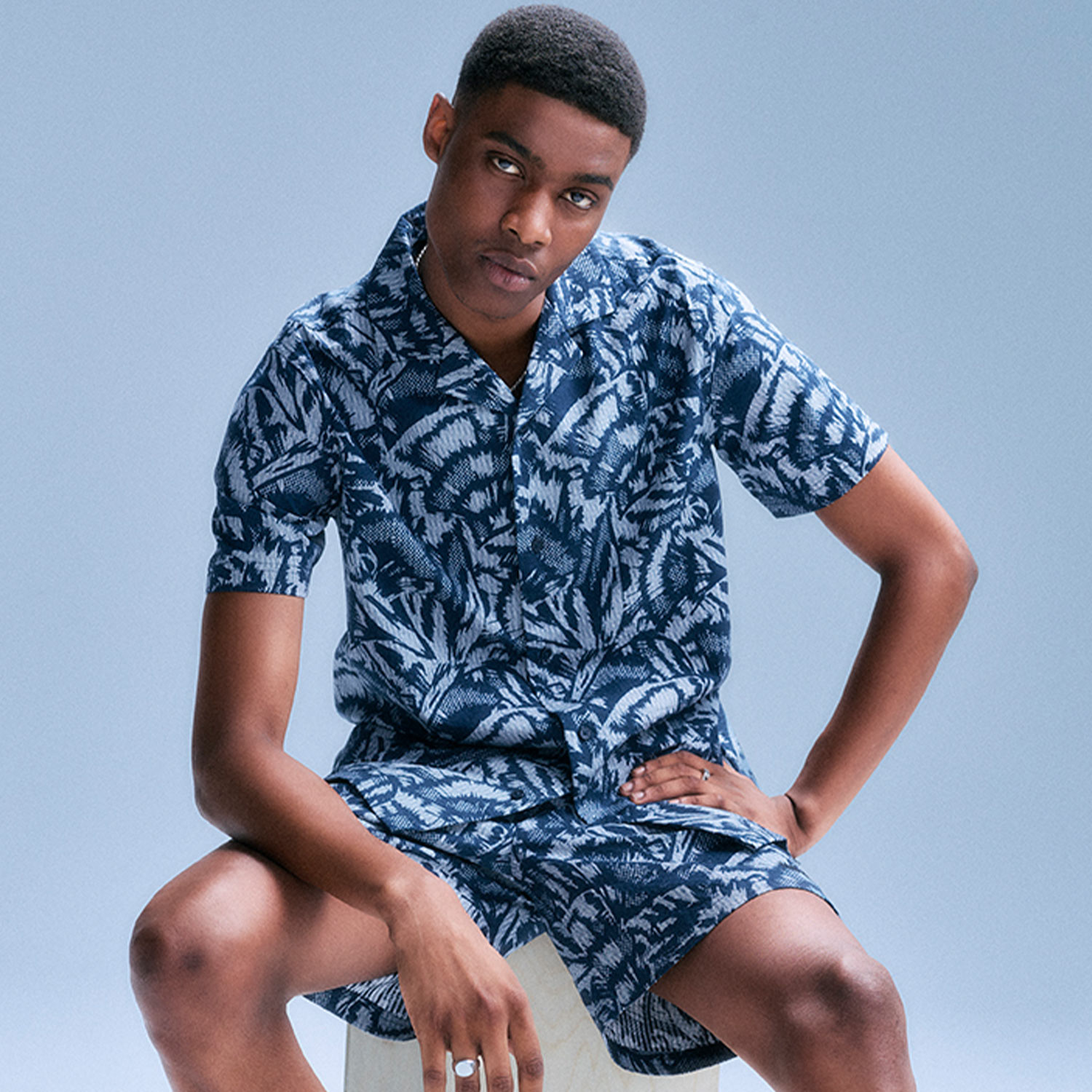Person seated on a stool wearing a blue patterned short-sleeve shirt and shorts, with a neutral expression.