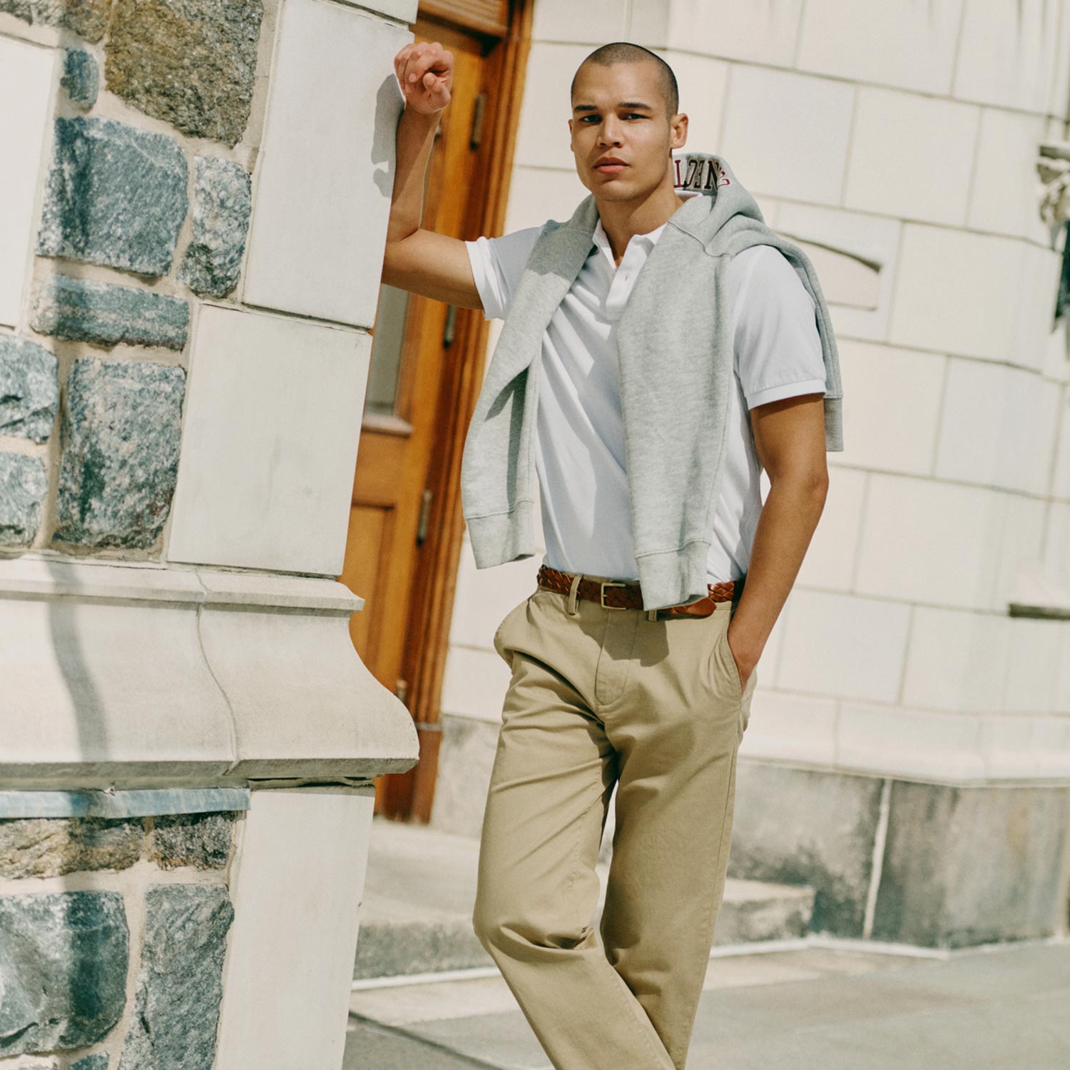 A person leans against a stone building, wearing a white polo, beige pants, and black shoes, with a sweater draped over their shoulders.