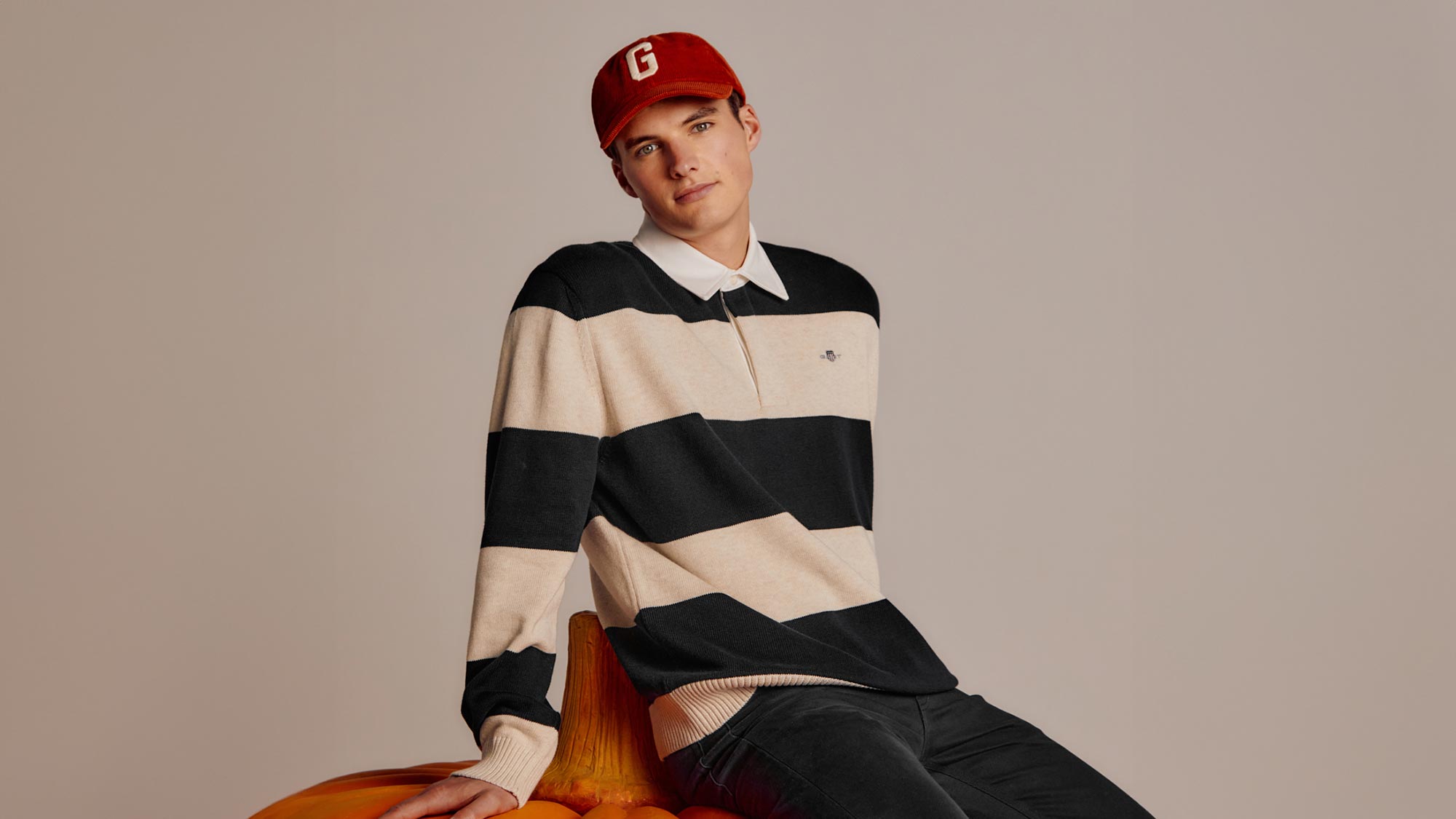 A person in a striped rugby shirt and red cap sits on a large pumpkin against a neutral background.