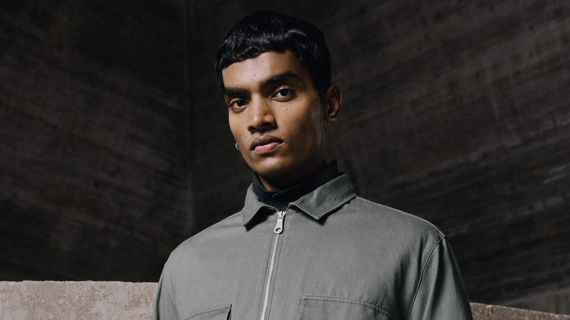 A person in a green jacket and dark pants poses in a concrete interior setting.