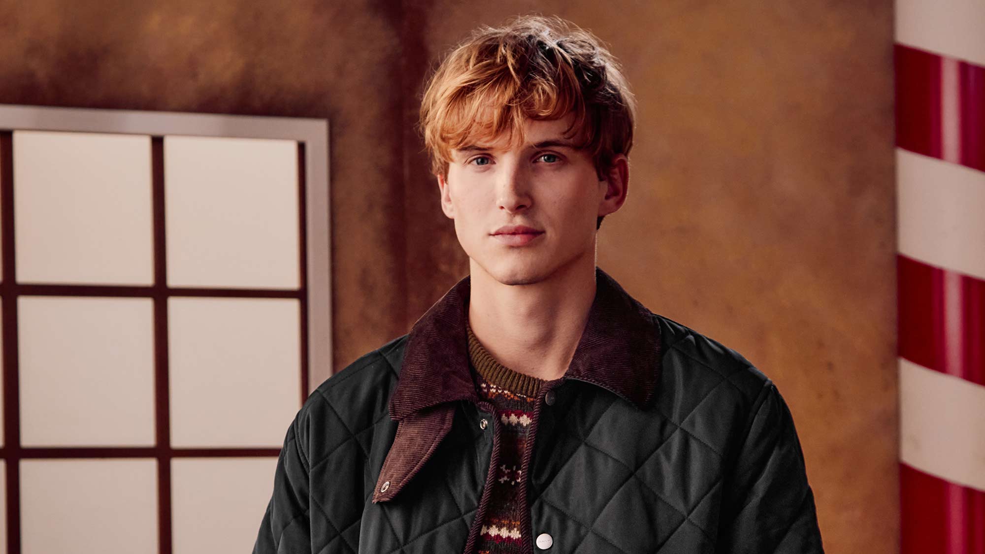 A person in a quilted jacket and patterned sweater stands in front of a festive gingerbread-themed backdrop.