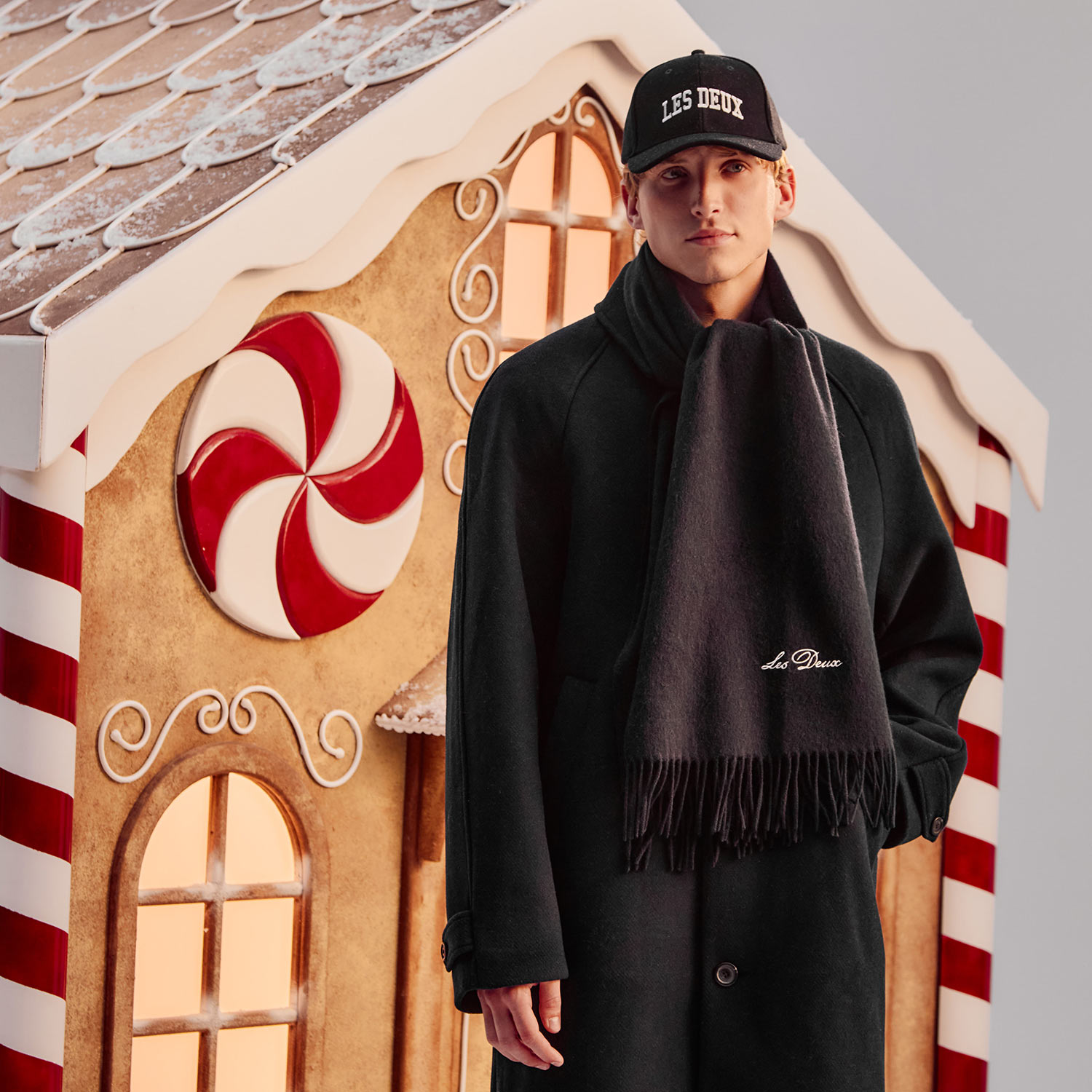 A person in a dark coat and cap stands in front of a gingerbread house with candy decorations.