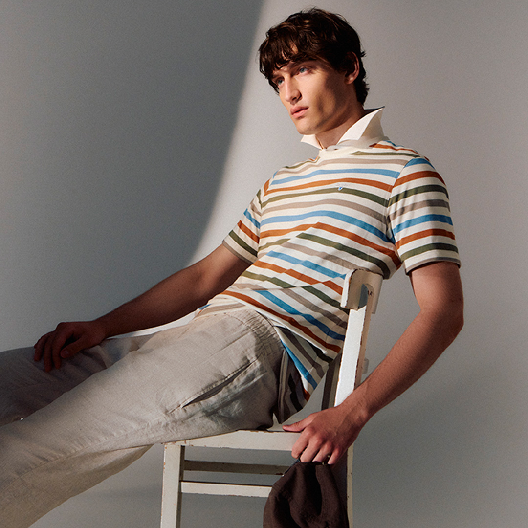 A person sits on a tilted chair, wearing a striped shirt and beige pants, holding a dark hat in one hand.