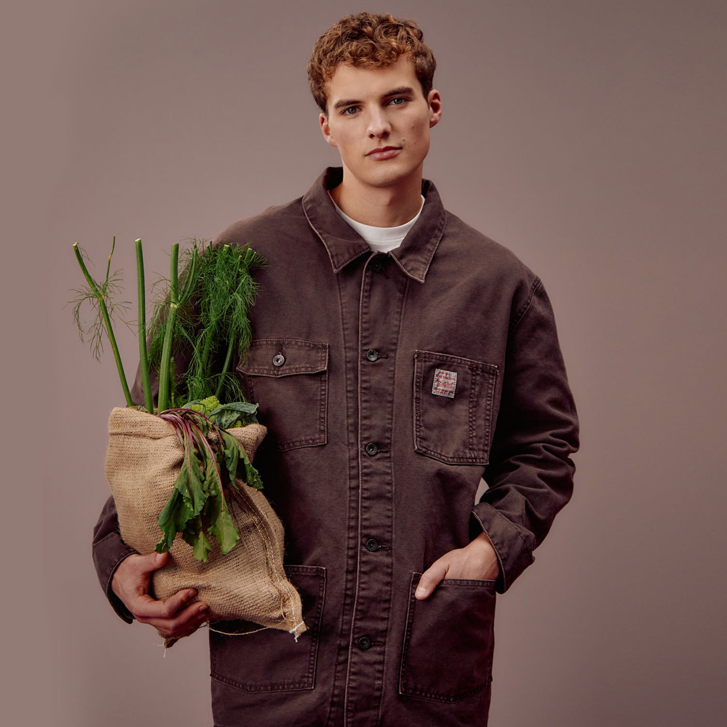 man in brown overshirt holding brown bag with green vegtable stalks sticking out