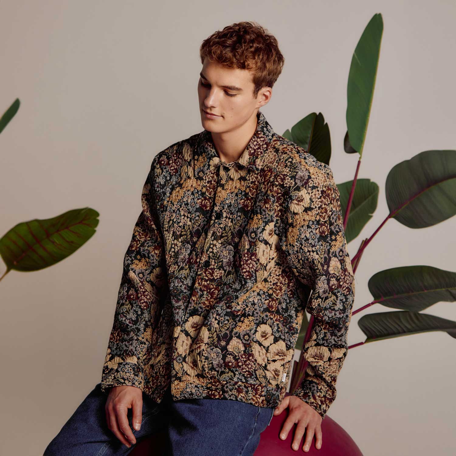 A person in a floral jacket sits on a large red orb with tropical leaves in the background.