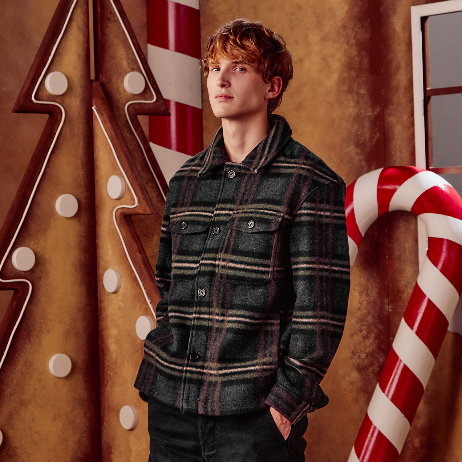 A person in a plaid jacket stands in a gingerbread-themed setting with candy cane decorations.