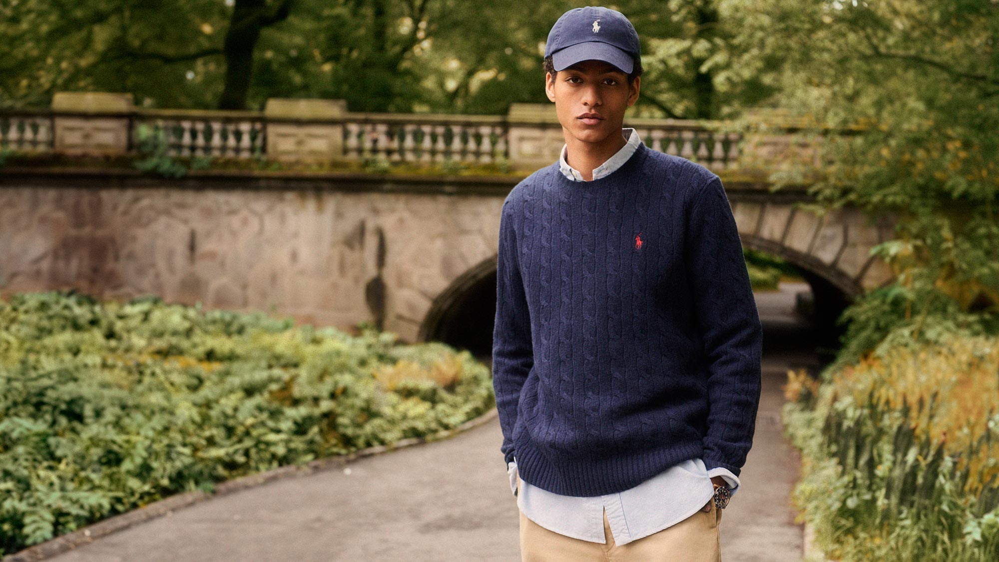 A person wearing a blue sweater and cap, standing on a path in a lush, green park with a stone bridge in the background.