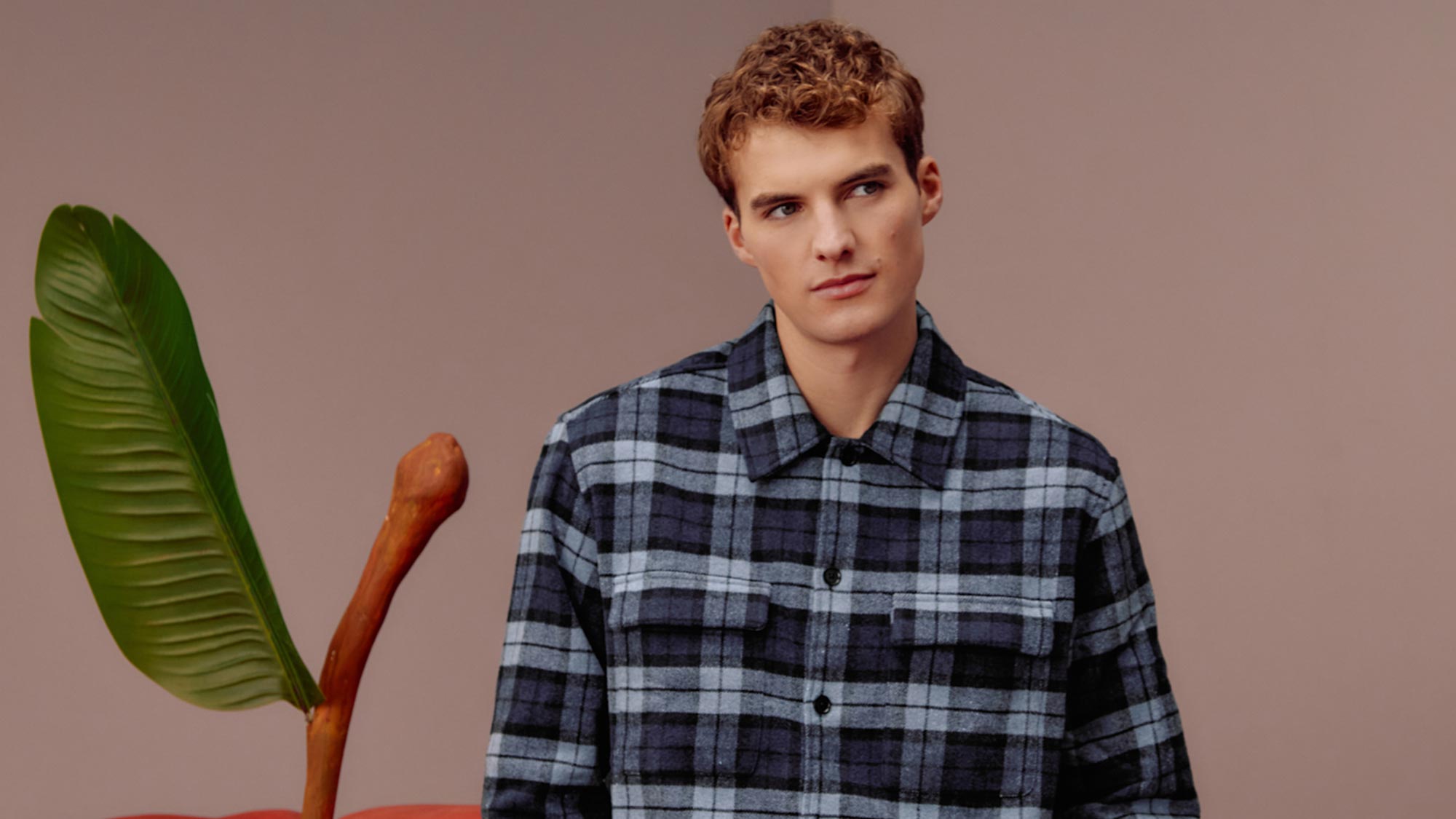 A person in a plaid shirt leans against a large, artificial apple with a leaf and stem in a minimalist setting.
