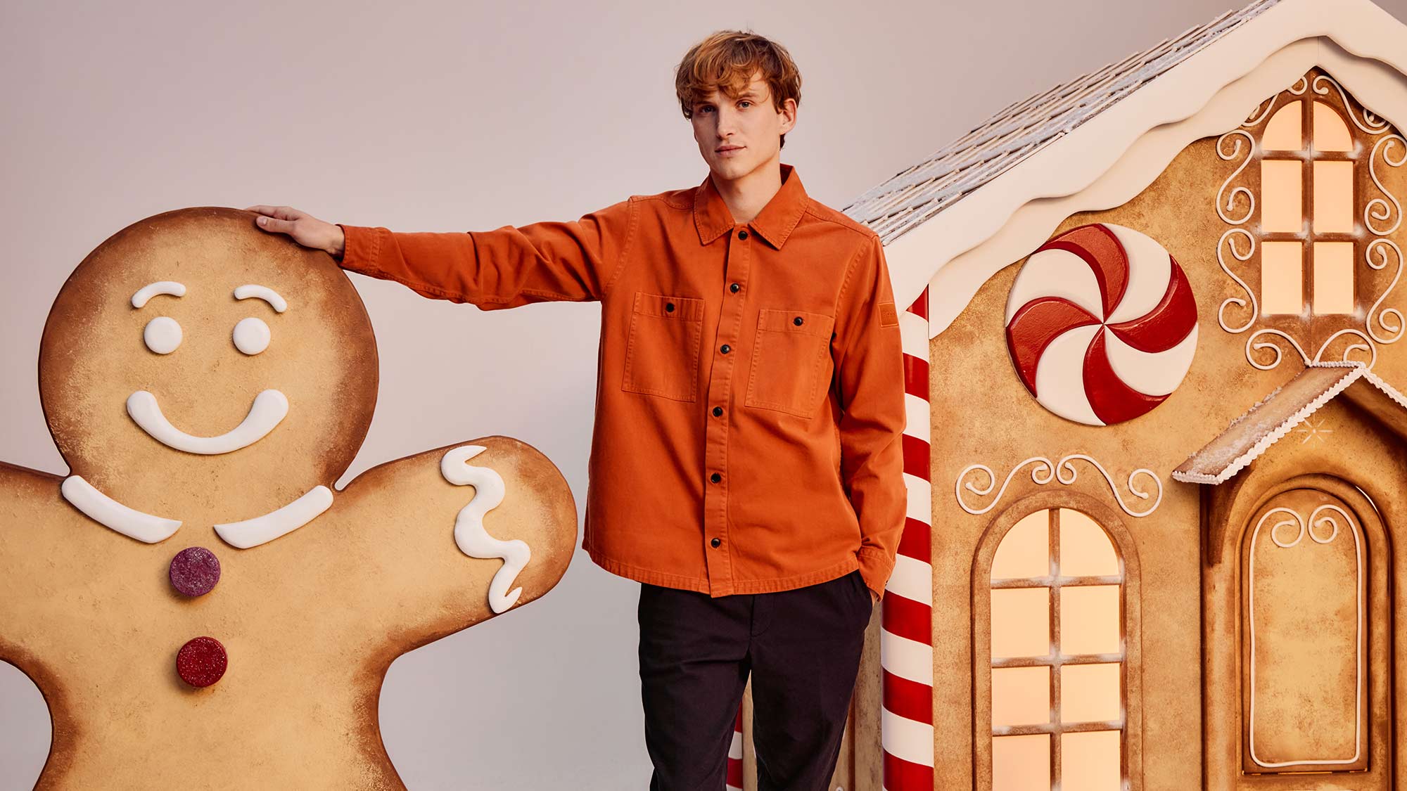 A person in an orange jacket stands next to a large gingerbread figure and house.