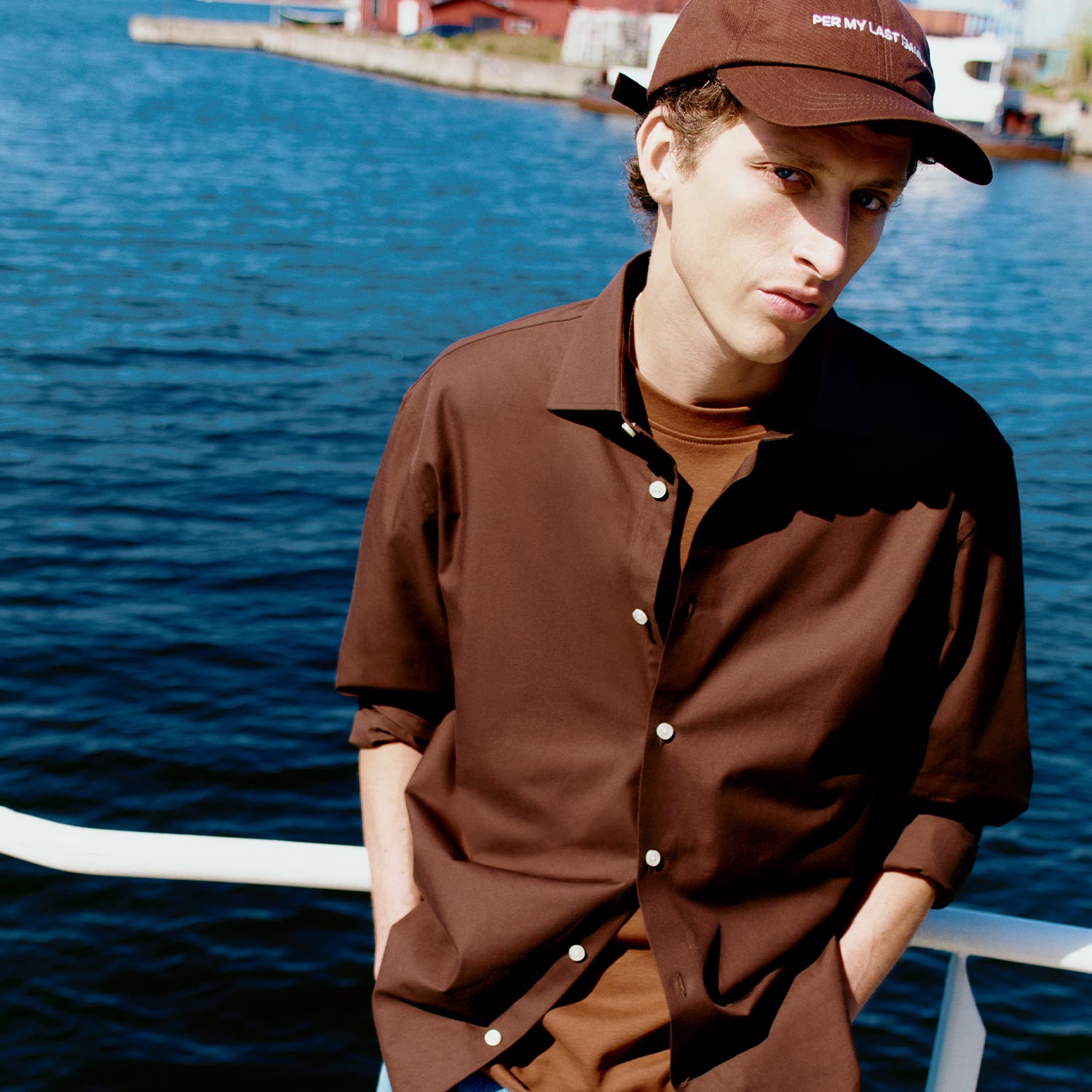 A person wearing a brown shirt and cap stands on a dock by the water.