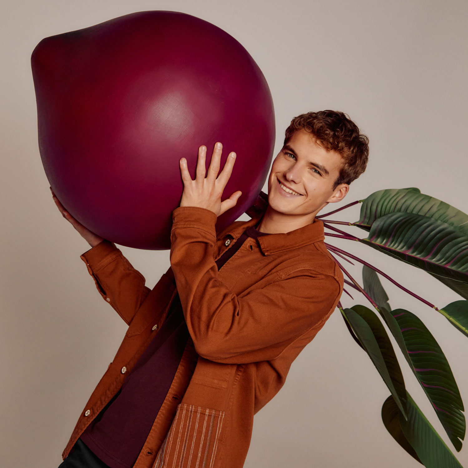 A person in a brown jacket holds a large, purple, pear-shaped object with green leaves in the background.