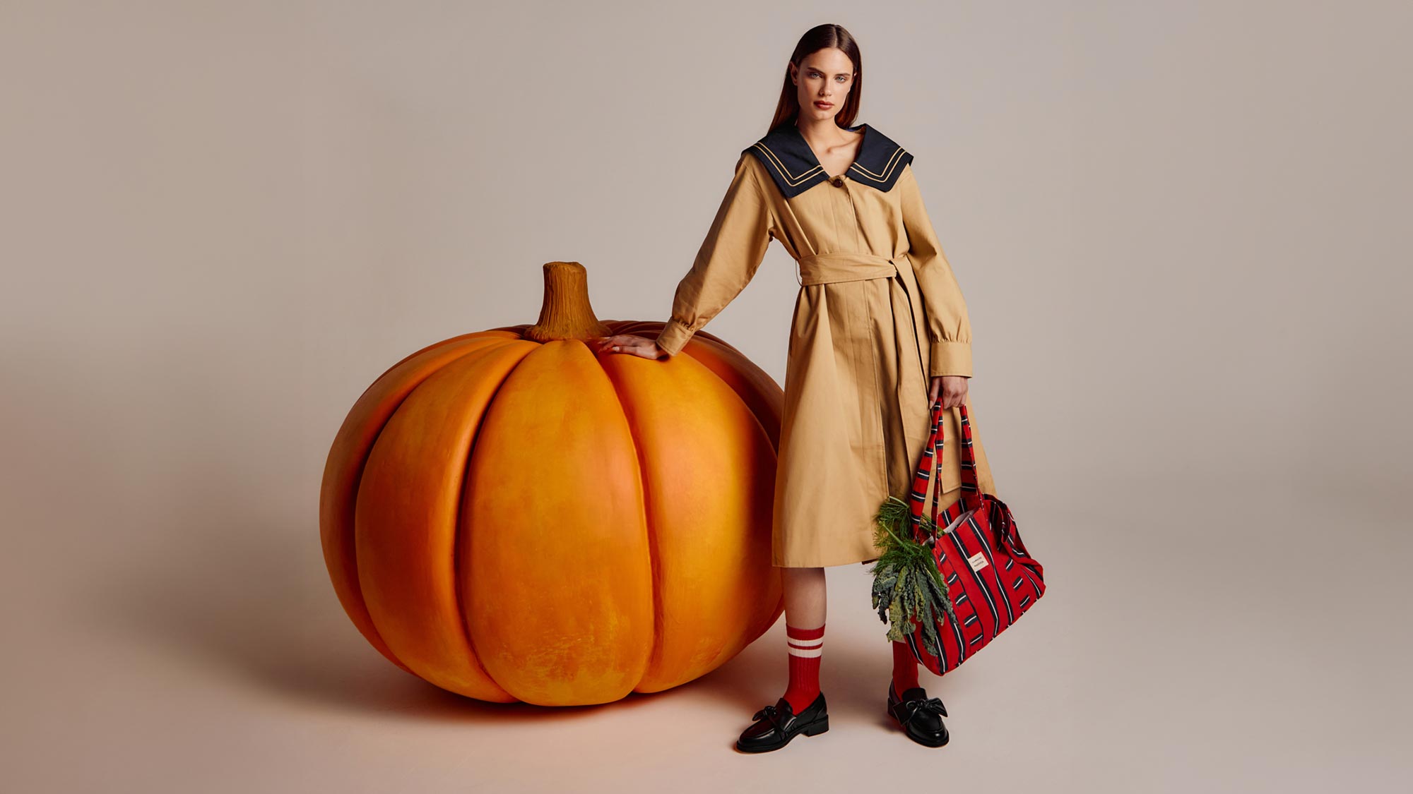 A person in a beige coat stands beside a large pumpkin, holding a plaid bag with greens. They wear red socks and black shoes.