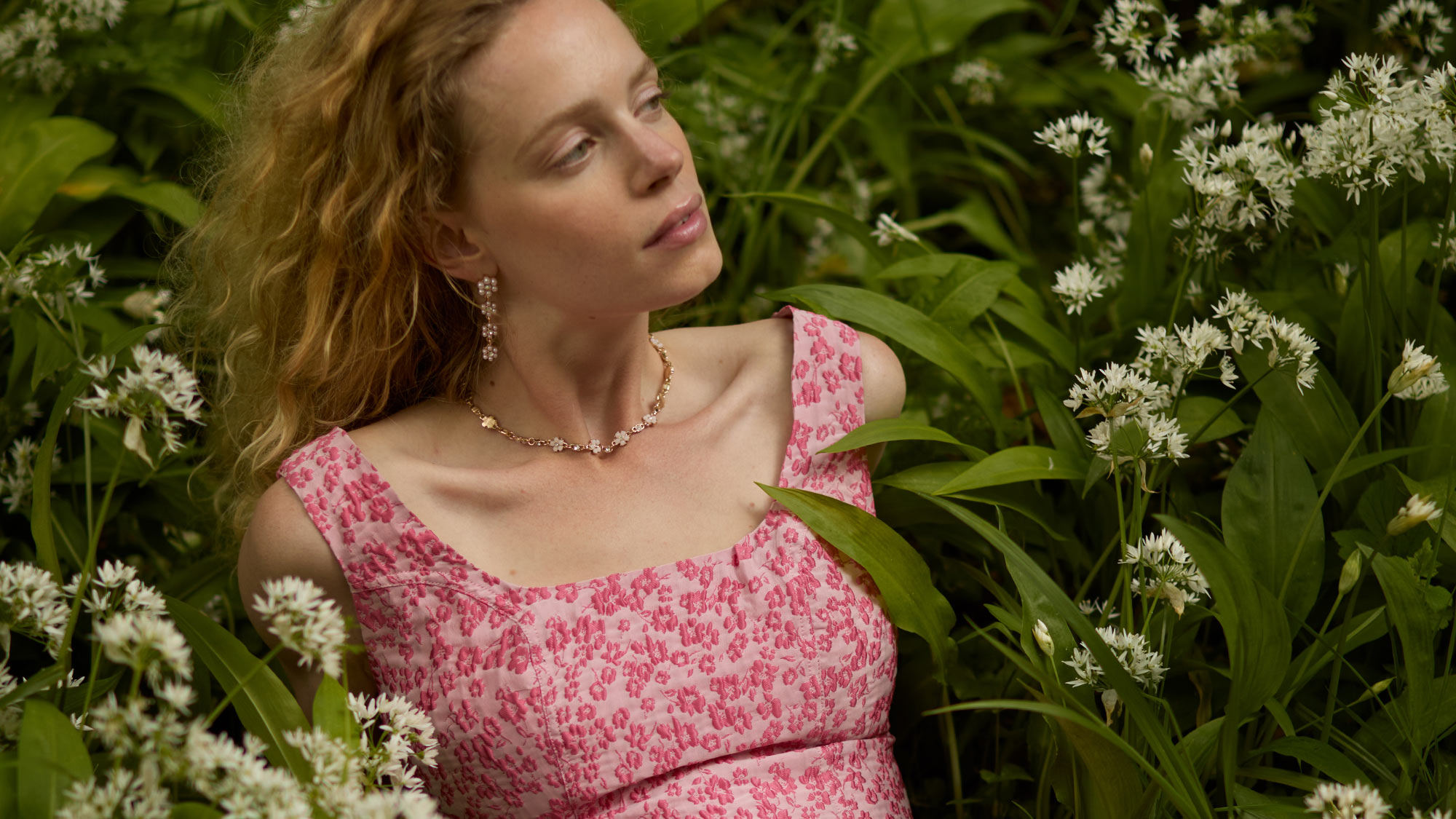 A woman in a pink floral dress lies relaxed among lush green plants and white flowers.
