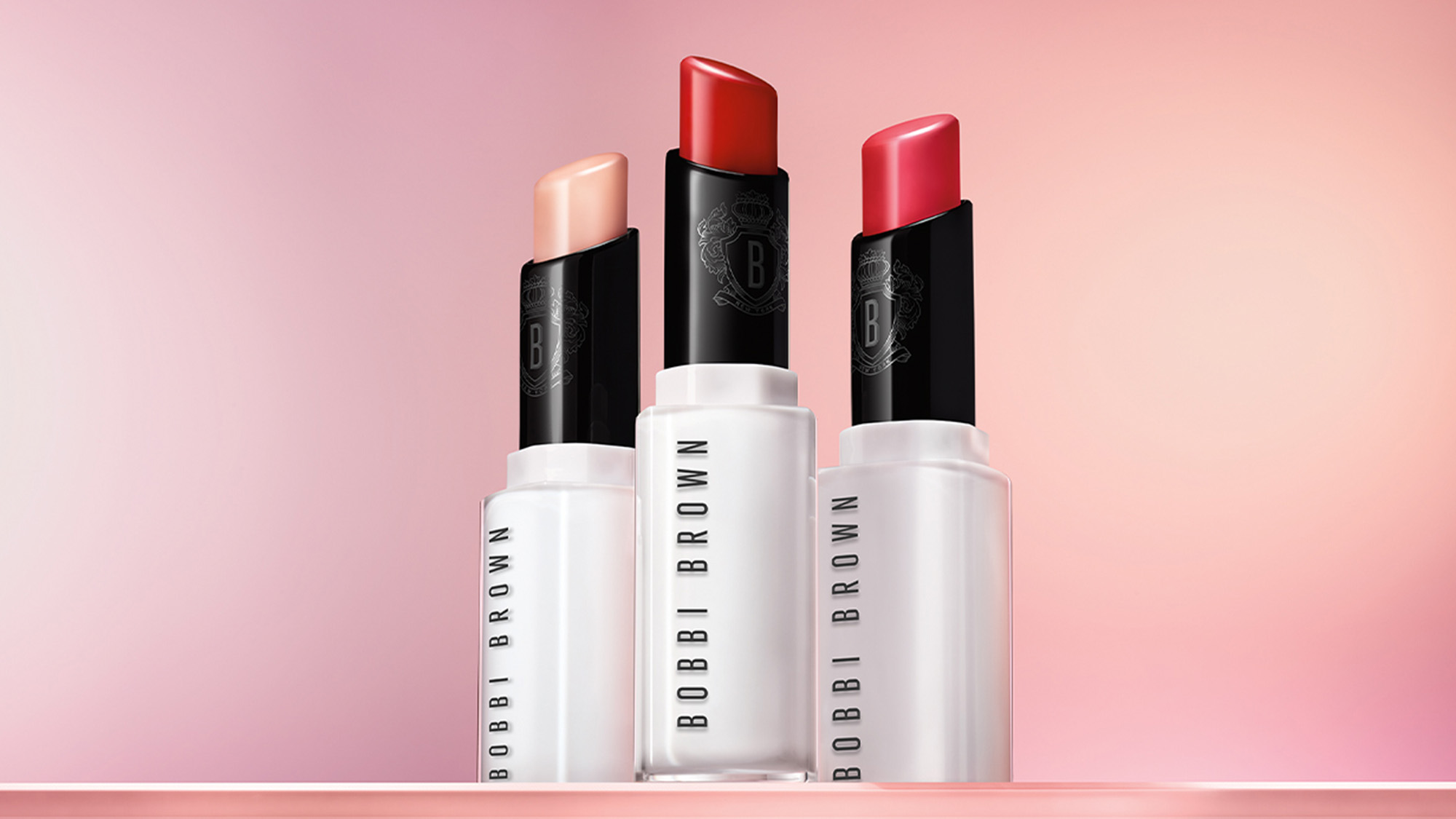 Three Bobbi Brown lipsticks in different shades on a pink gradient background.
