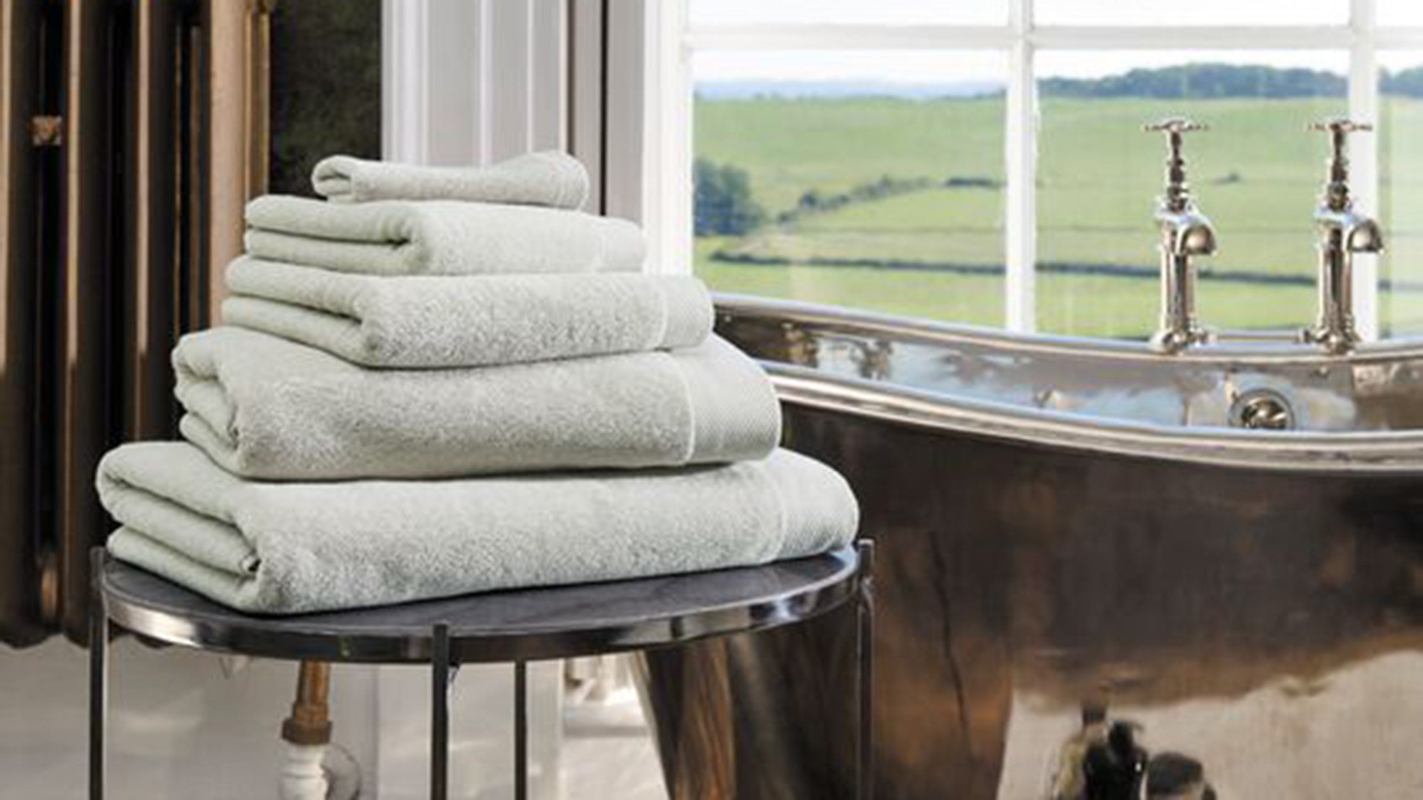 Stacked towels on a round table beside a shiny bathtub with countryside view.