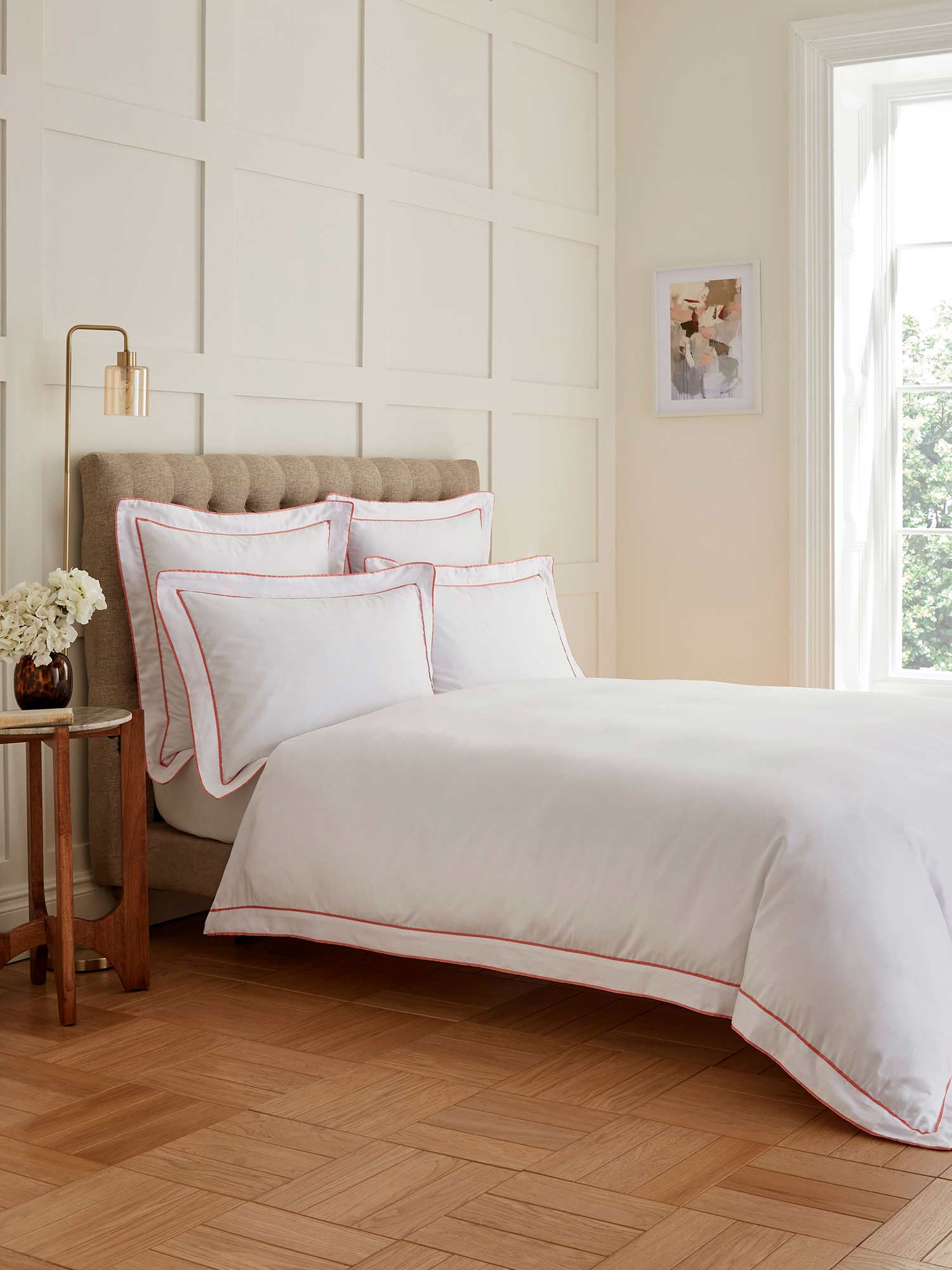 Bright bedroom with a neatly made bed, white and pink bedding, and a large window. A side table holds flowers beside the bed.