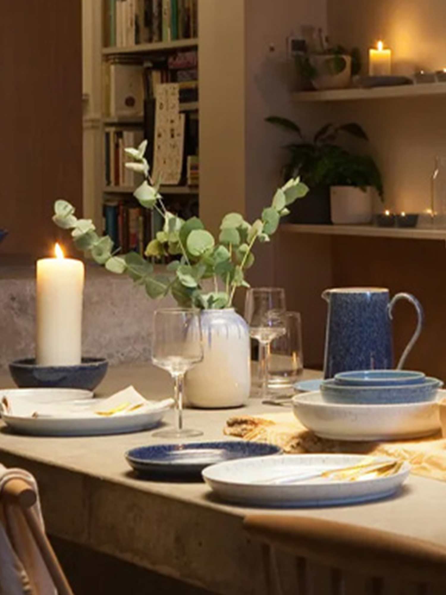 A cozy dining setup with blue plates, glassware, and potted plants on a wooden table, with candles and shelves in the background.