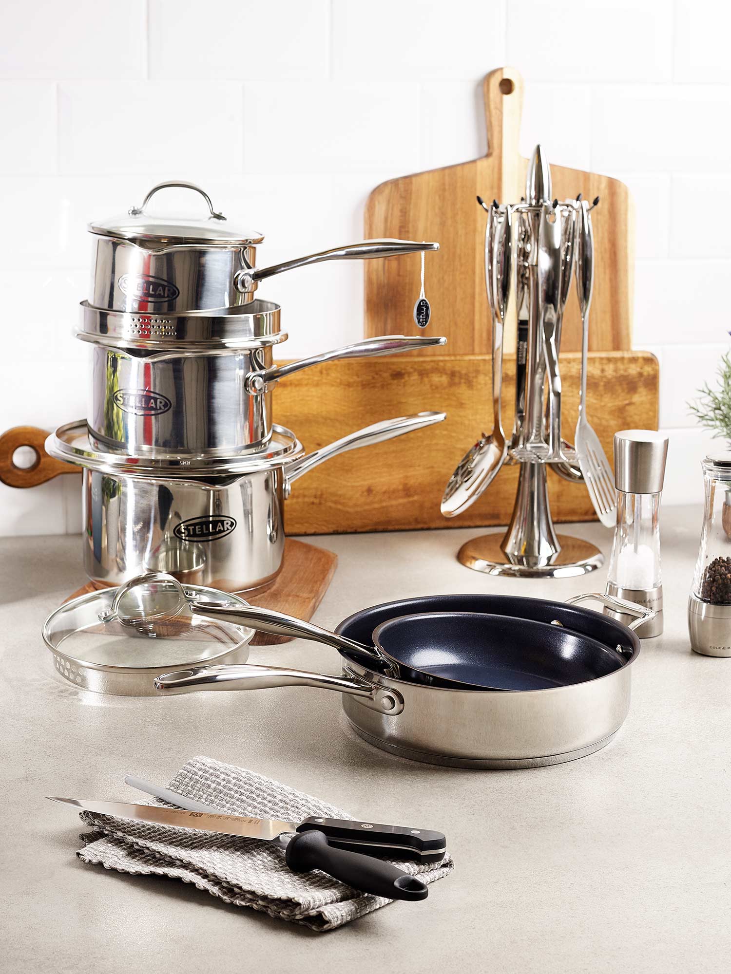 A set of stainless steel cookware, cutlery, and utensils on a kitchen counter with a wooden cutting board in the background.