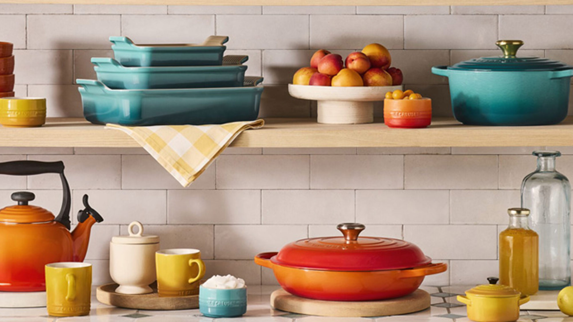 Colourful kitchen cookware and dining accessories on white brick shelving, including teal casserole dishes, orange kettle, and yellow mugs