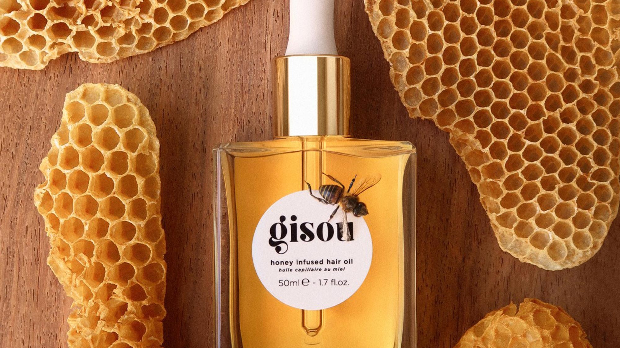 A bottle of honey-infused hair oil by Gisou is surrounded by honeycombs on a wooden surface.