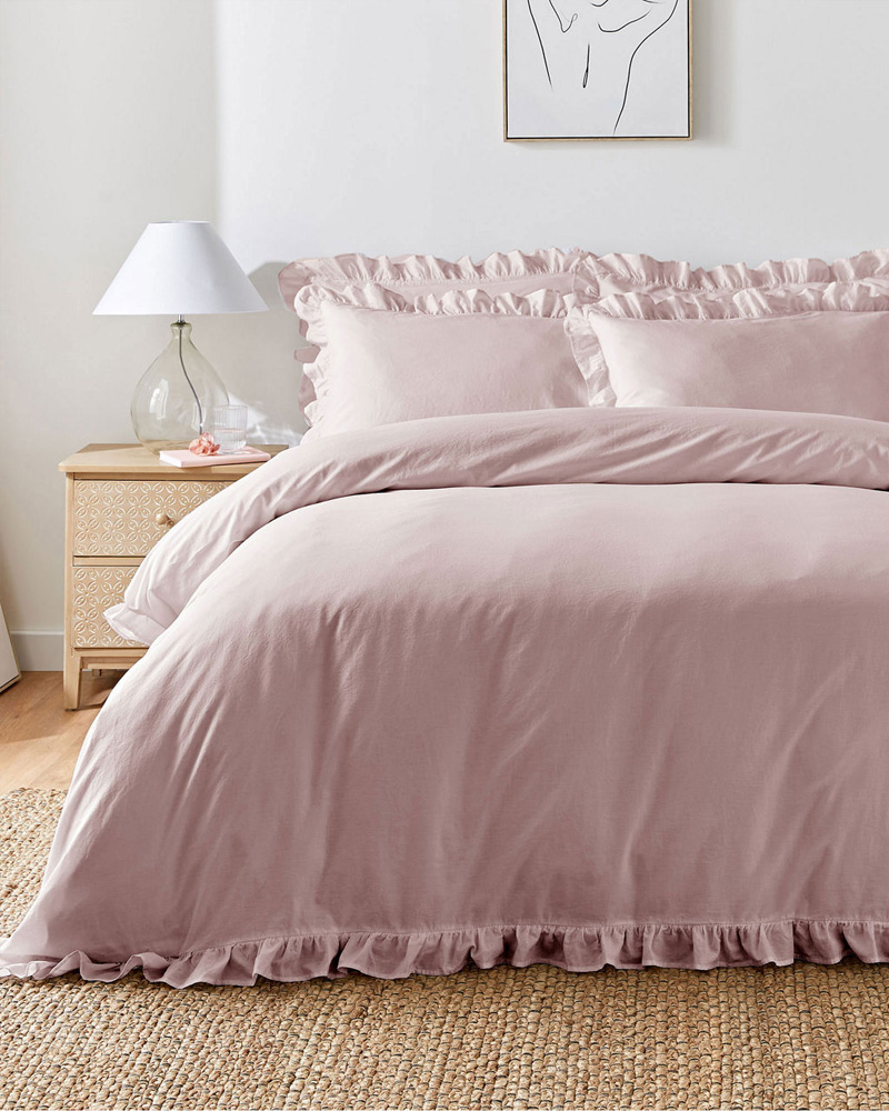 A neatly made bed with pink ruffled bedding stands beside a wooden nightstand with a lamp. A simple line drawing hangs above.
