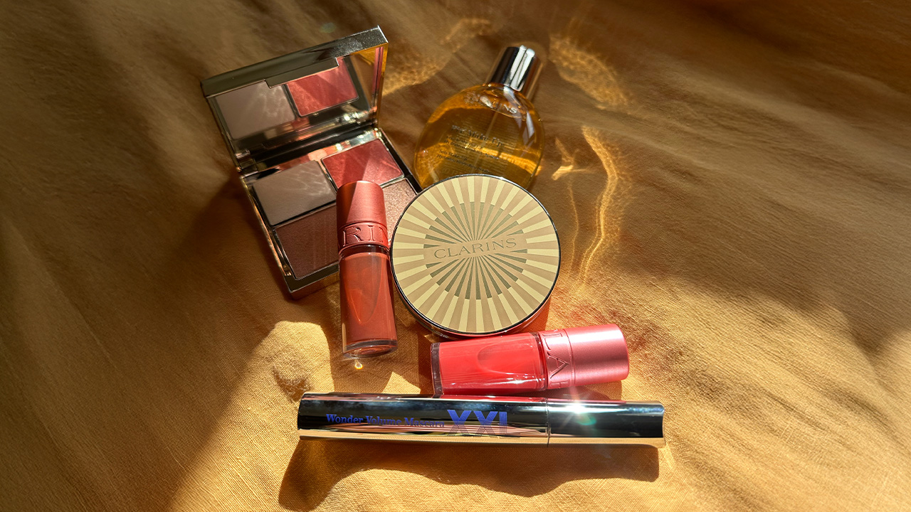 Clarins makeup products, including a compact, lip glosses, and a mascara, arranged on a sunlit golden fabric.