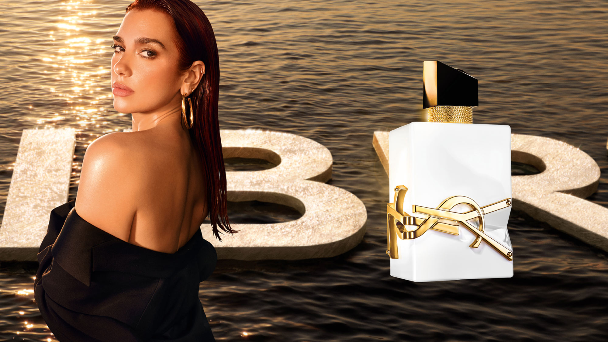 Dua Lipa poses with a YSL Libre L'eau Nue perfume bottle, set against a shimmering, golden backdrop.