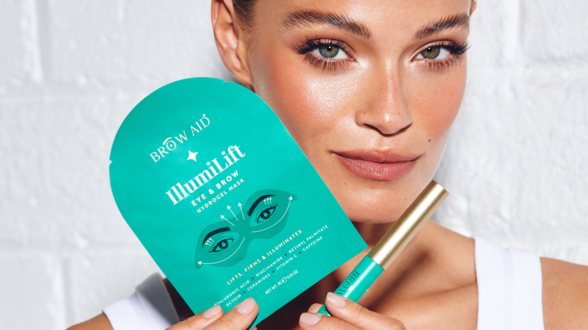 A person holds a teal brow and eye makeup product package and a tube against a white background.