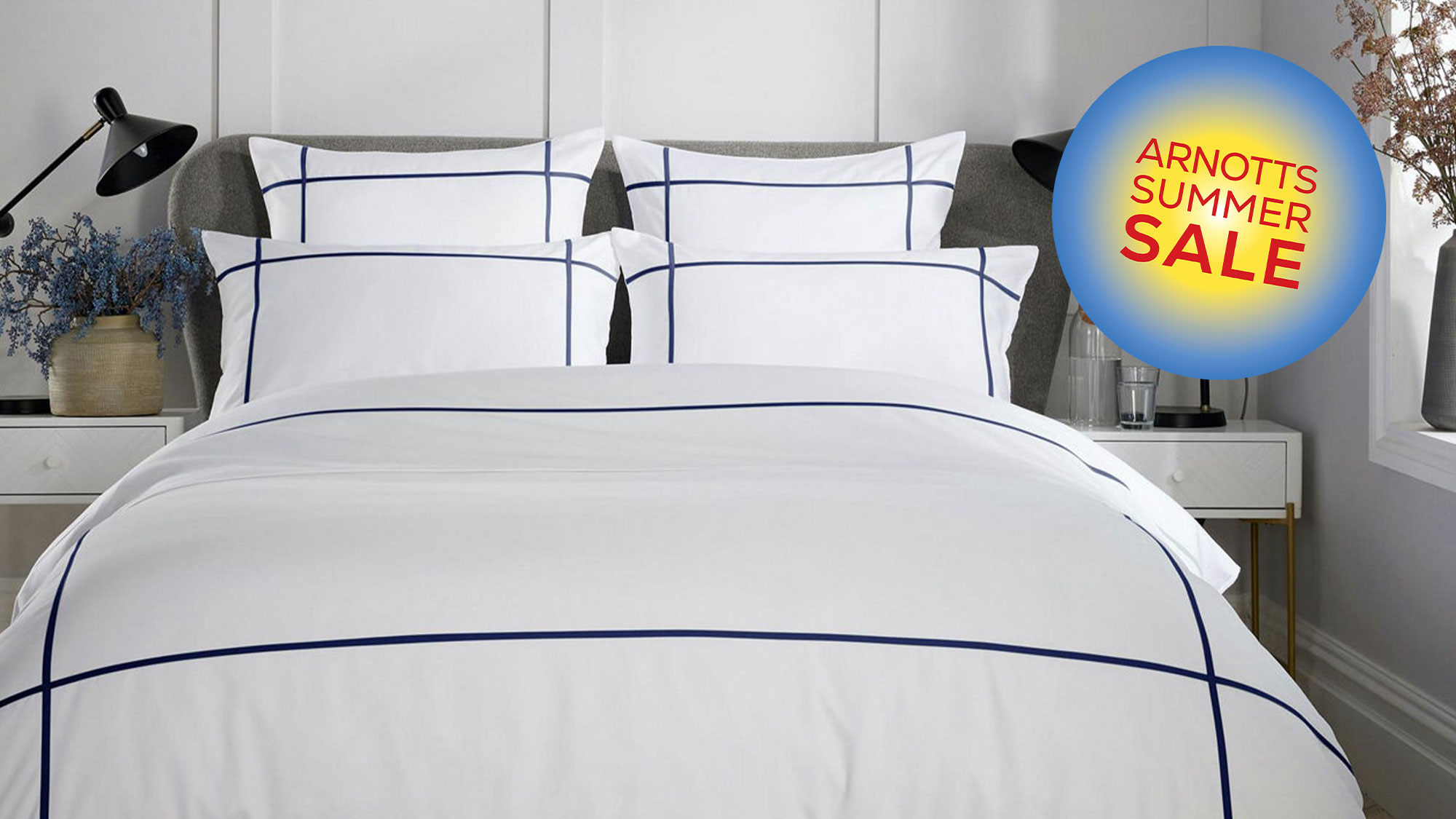 Modern bedroom with a neatly made bed, white and navy geometric bedspread, black lamps, and decorative plants.