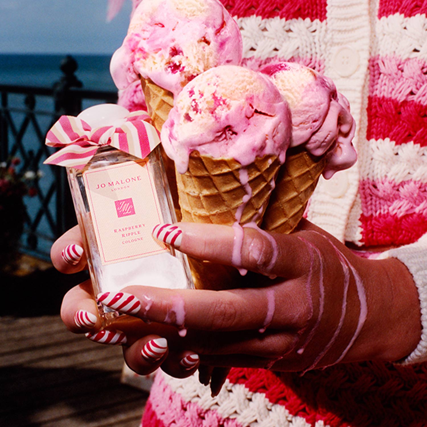 Person holding Jo Malone London perfume with raspberry ripple ice cream cones against a seaside backdrop.