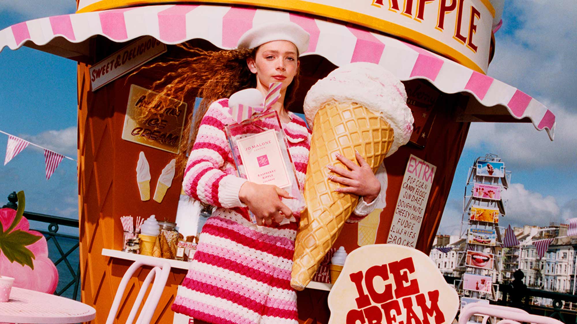 Person holding Jo Malone London perfume with raspberry ripple ice cream cones against a seaside backdrop.