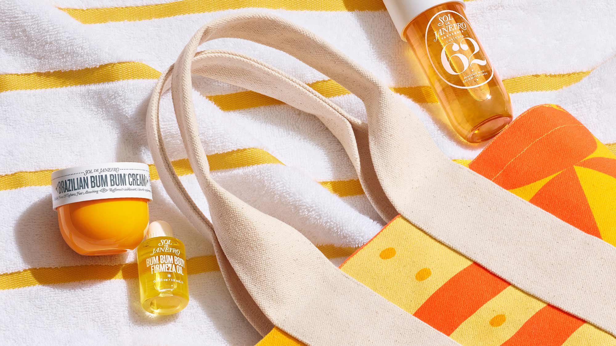 Sol de Janeiro products, including Brazilian Bum Bum Cream and body oils, are arranged on a striped towel next to a colorful tote bag.