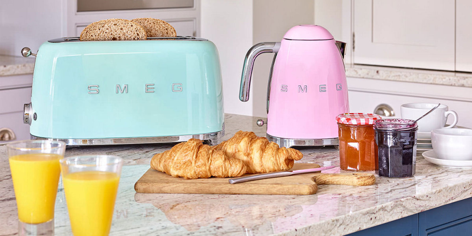 Retro-style SMEG toaster and kettle in a kitchen with croissants, juice, and jams on the countertop.