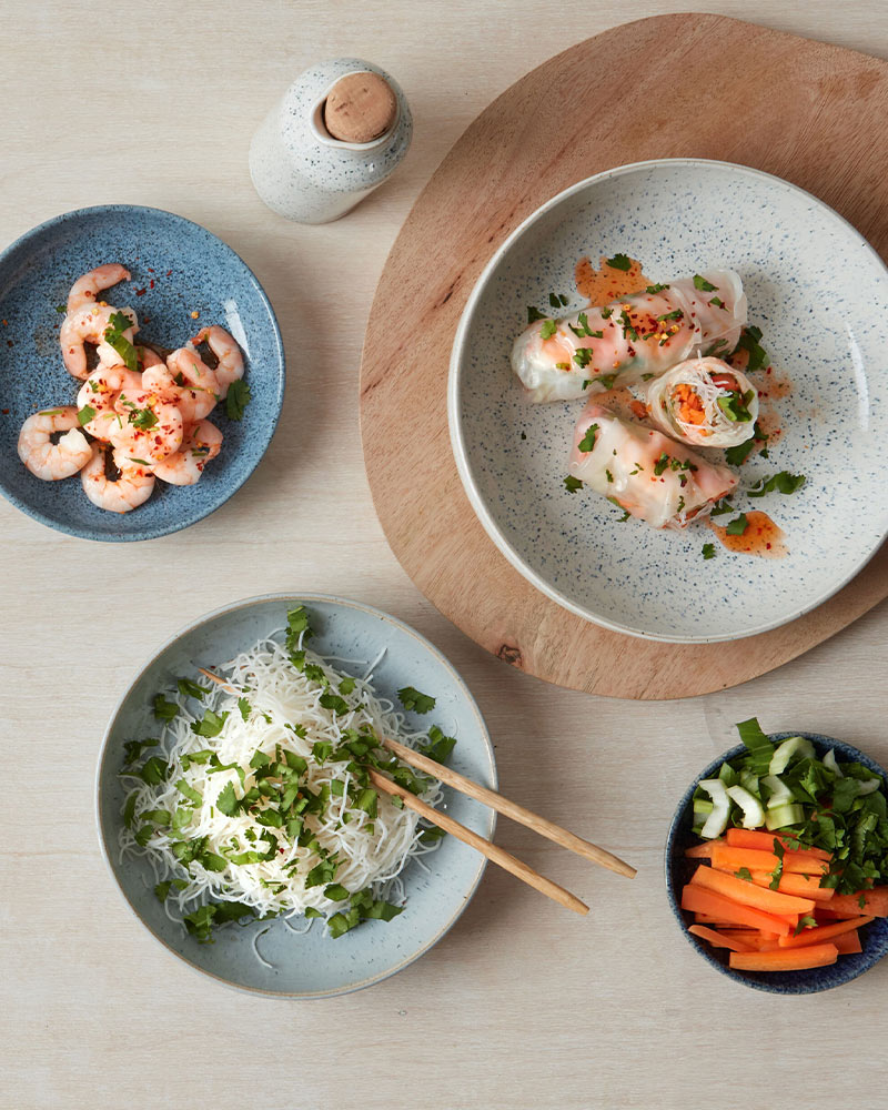 Plates of shrimp, spring rolls, rice noodles, and vegetables arranged on a wooden board with chopsticks and a small bottle.