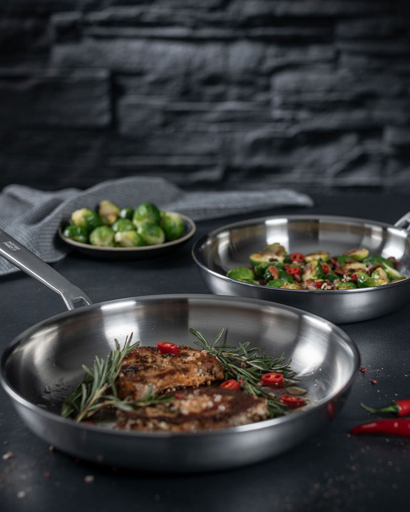 Two stainless steel pans with cooked meat, rosemary, and Brussels sprouts. A plate of uncooked Brussels sprouts in the background.