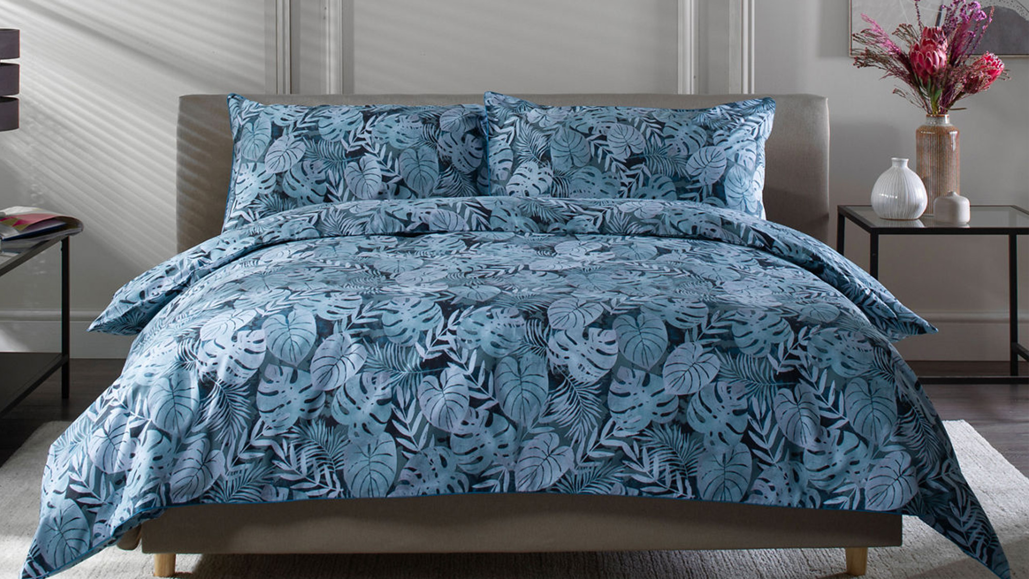A bed with blue leaf-patterned bedding in a modern, minimalistic room. A side table with a vase and artwork on the wall completes the scene.