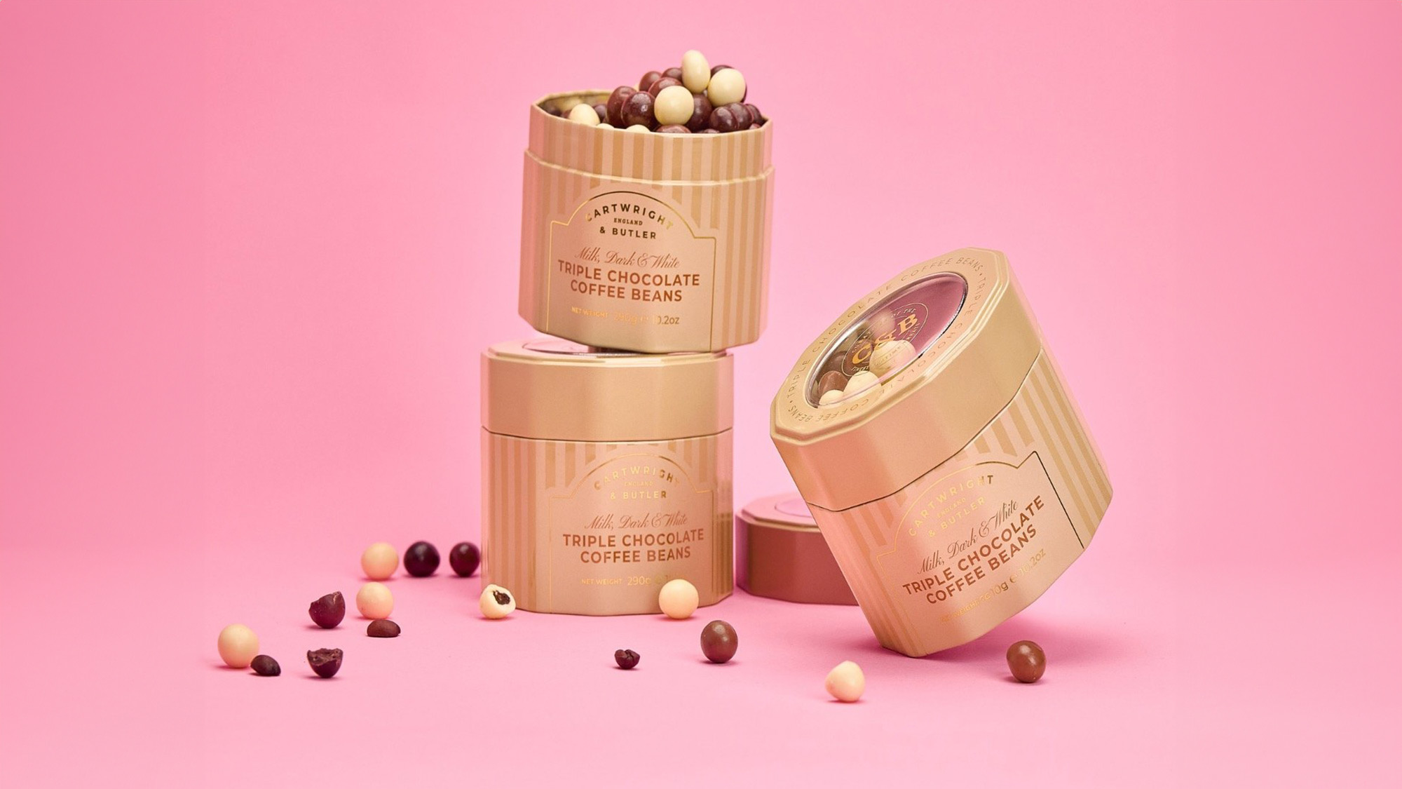 Three containers of triple chocolate coffee beans on a pink background, with some beans scattered around.