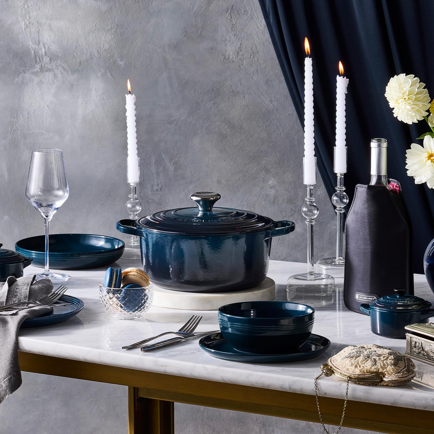 Elegant table setting with dark blue crockery, lit candles, a wine bottle, and white flowers, against a gray backdrop with navy curtains.