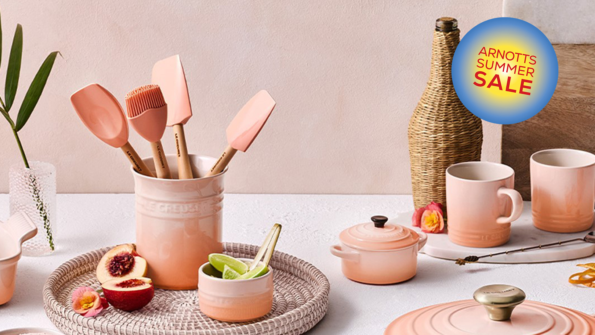 A collection of peach-colored kitchenware on a white surface, featuring spatulas, pots, a glass, and a cutting board with decorative items.