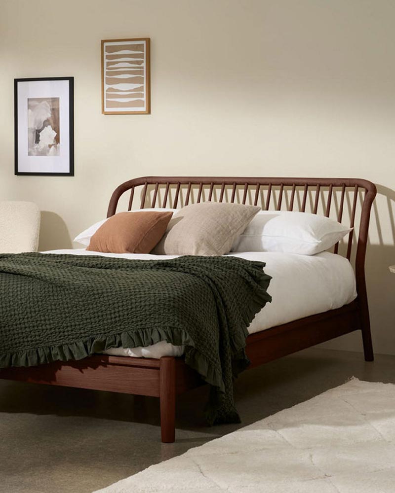 A cozy bedroom with a wooden bed, green blanket, assorted pillows, beige wall, and framed art.