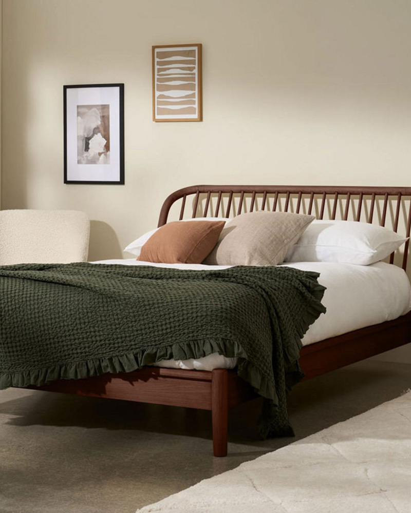 A cozy bedroom with a wooden bed, green blanket, assorted pillows, beige wall, and framed art.