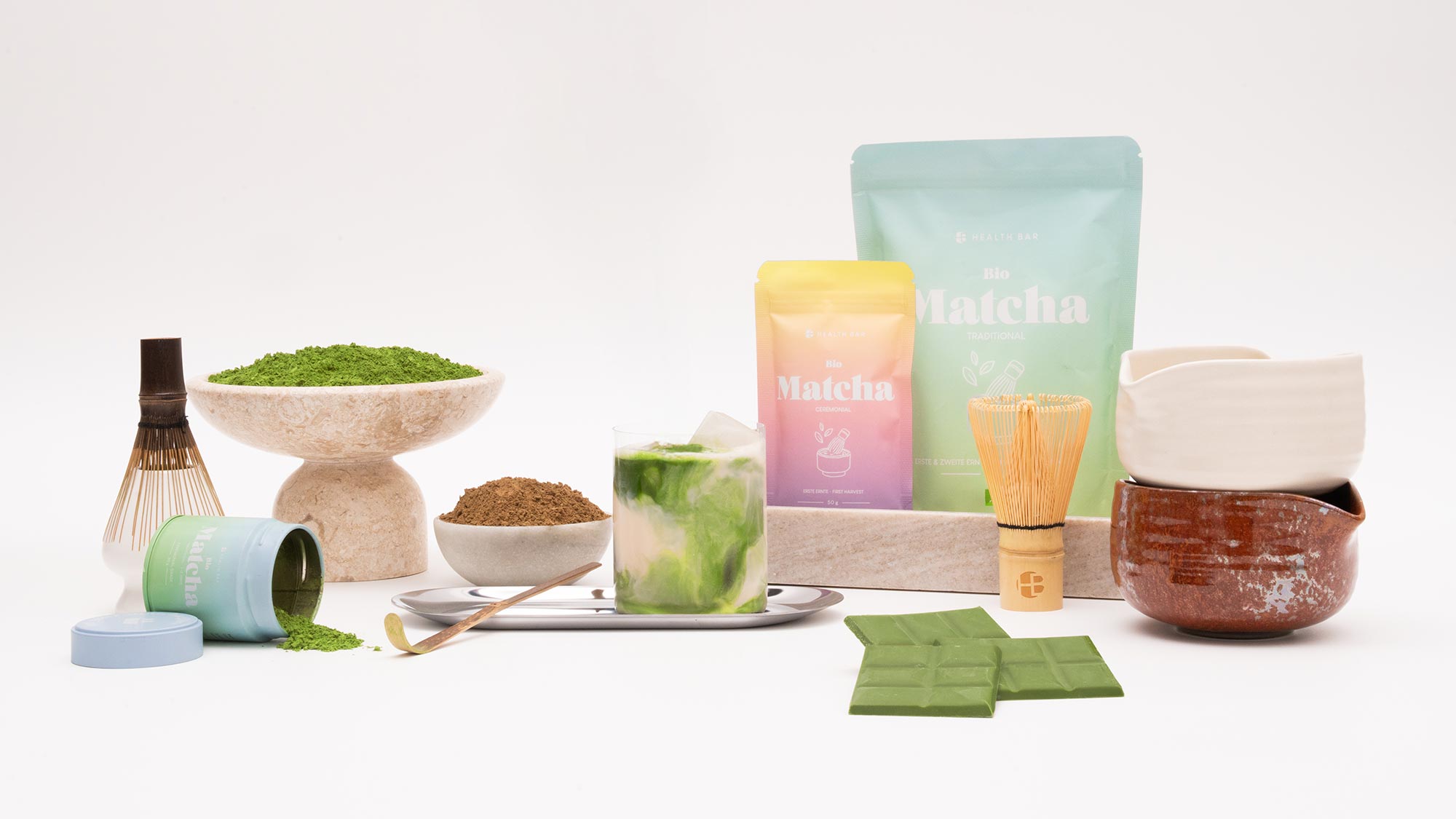 A variety of matcha tea products, including powdered tea, a whisk, and a cup, arranged on a white surface.