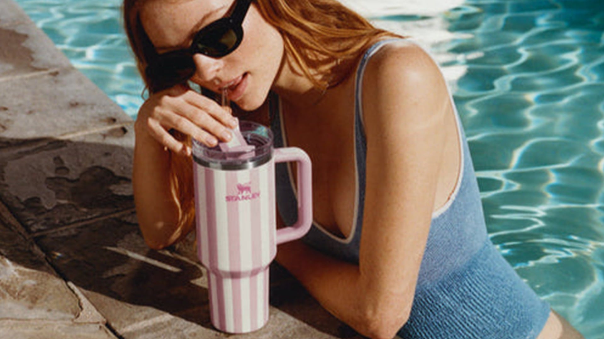 A person in sunglasses and a blue swimsuit drinks from a large striped cup by a pool.