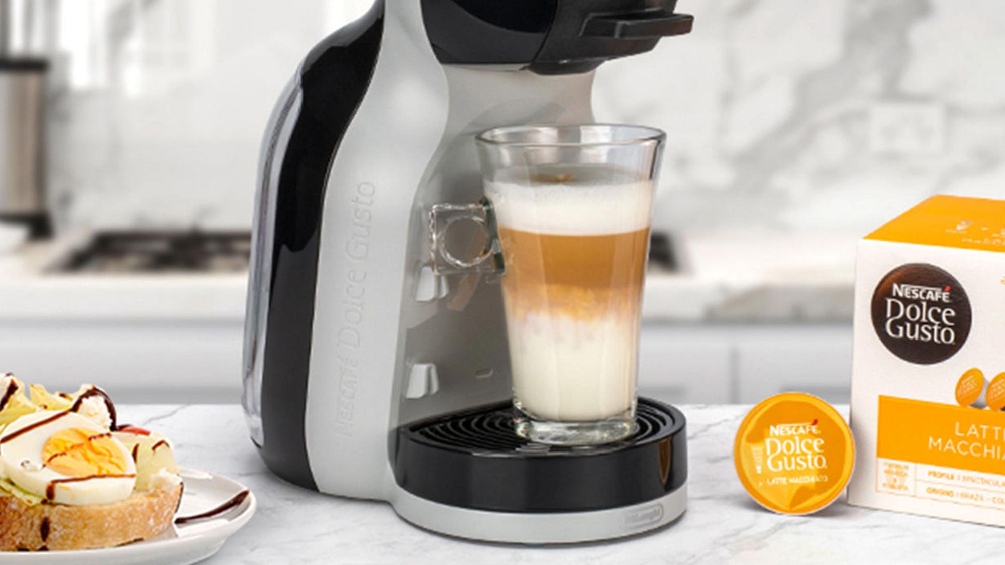 A coffee machine with a latte macchiato in a glass, a plate with sandwiches, and a box of Dolce Gusto capsules on a kitchen counter.