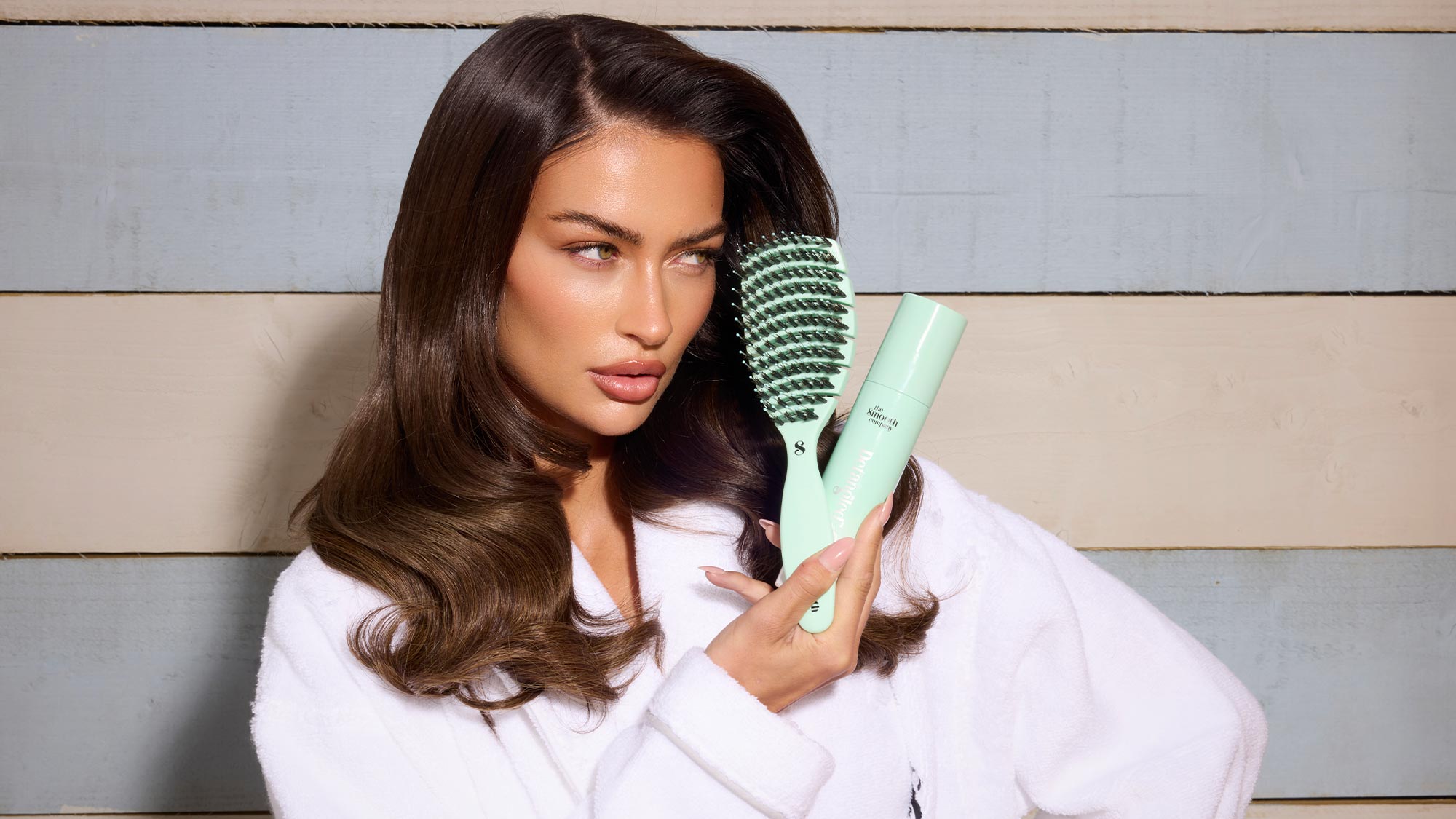 A person in a white robe uses a mint-green hairbrush. The background features wooden planks in neutral tones.