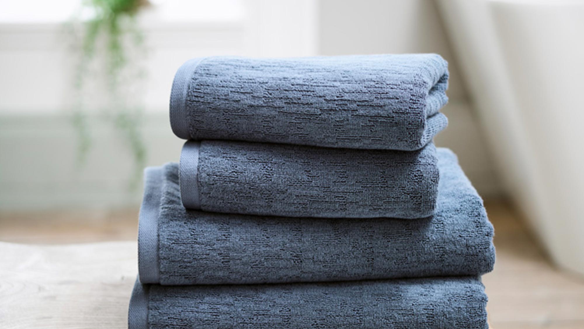 A stack of blue towels on a wooden bench in a bright bathroom setting.
