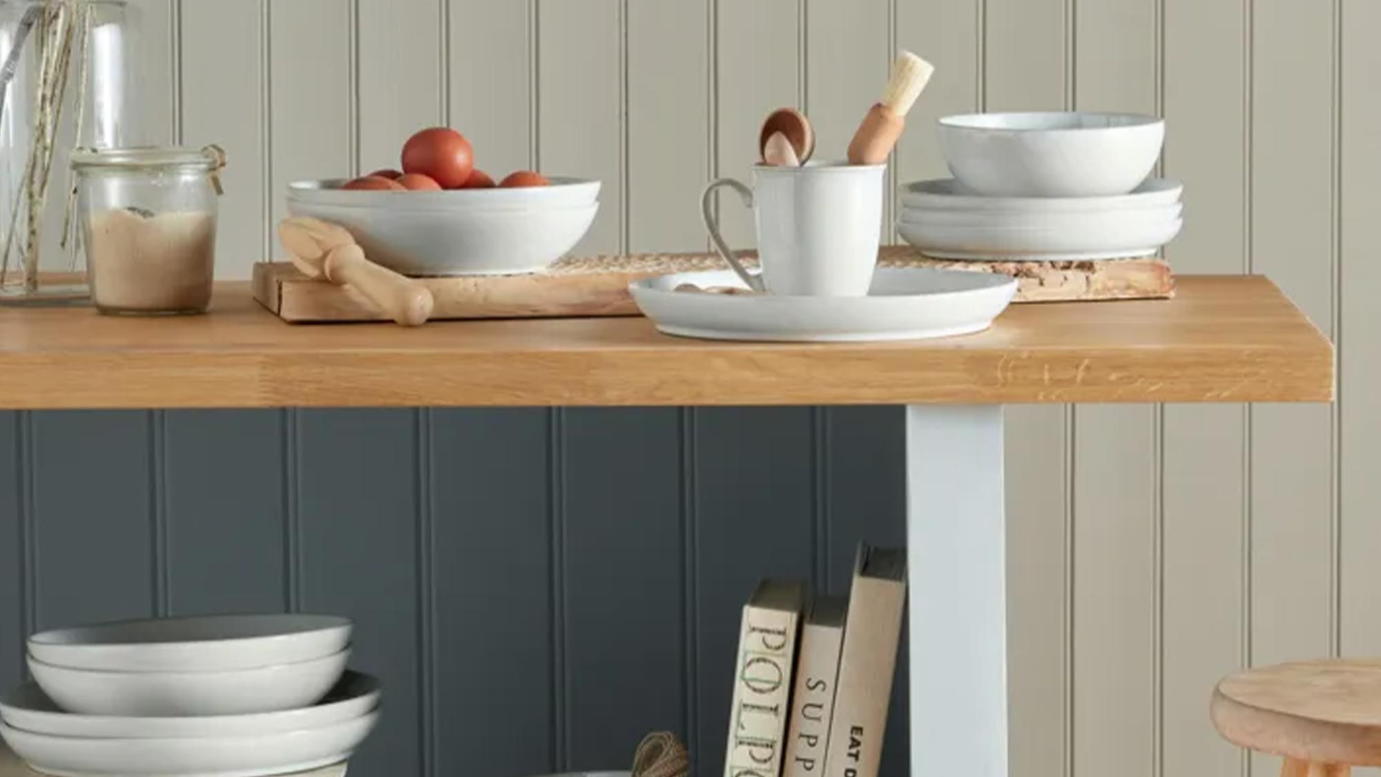 A kitchen scene with wooden shelves displaying bowls, plates, books, a cutting board with tomatoes, and a vase of dried flowers.