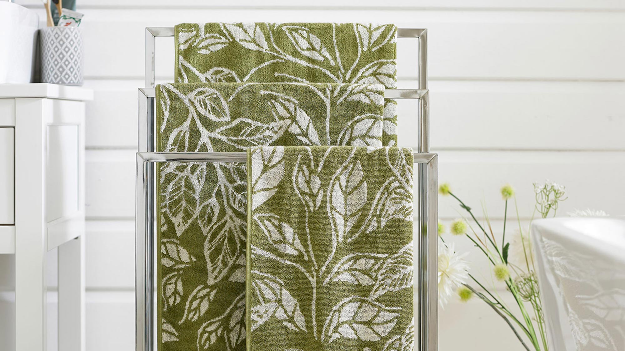 Green towels with white leaf patterns on a metal towel rack in a bright room with white decor.