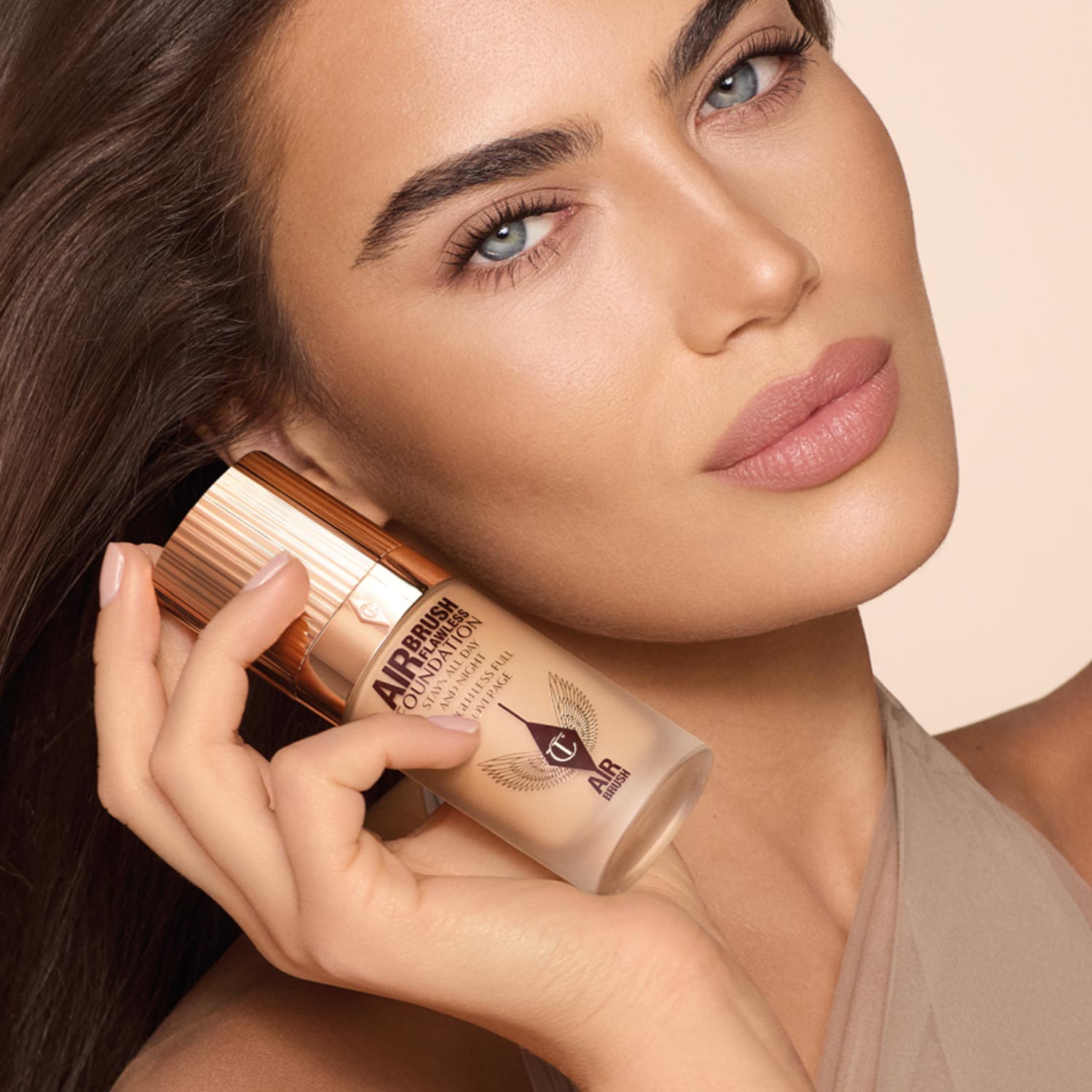 A woman holds a bottle of foundation on the left. On the right, three cosmetic products with shiny packaging and wing-like designs are displayed.